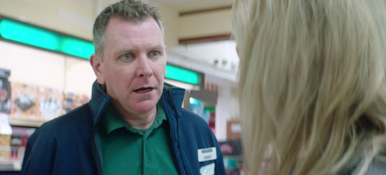 A Good Woman Is Hard To Find (2019) movie still: medium — The aisle of a brightly lit convenience store contains retail shelves in the background. A…