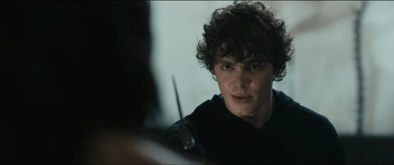 Scream VI (2023) movie still: medium — An interior space is dimly lit with cool tones. A young man with dark curly hair looks toward another person…