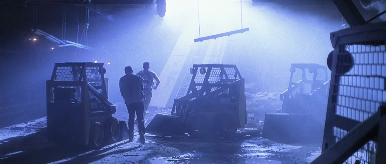 Die Hard with a Vengeance (1995) movie still: wide — A dimly lit indoor space filled with construction debris and thick atmospheric haze. Two men…
