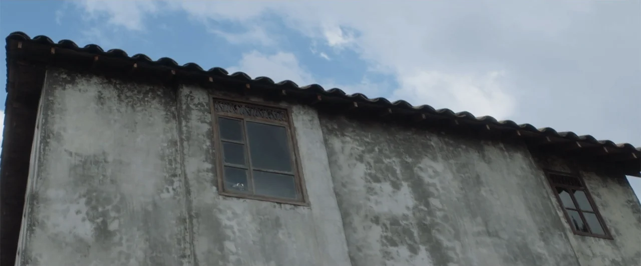 My Neighbor Adolf (2022) movie still: wide — A low-angle view shows the weathered concrete exterior wall of a two-story building. Two wooden-framed…
