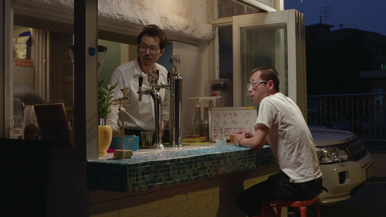 Yourself and Yours (2016) movie still: medium — An open-air bar counter with blue tiled edges sits on a dark city street. A man sits at the counter…