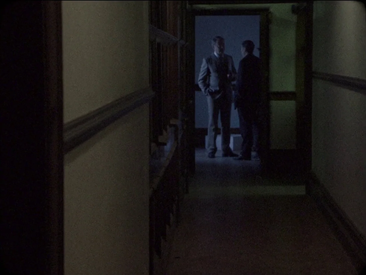 The Devil’s Doorway (2018) movie still: wide — A dark, narrow corridor with wood trim leads toward a brighter, illuminated doorway. Two men stand in…