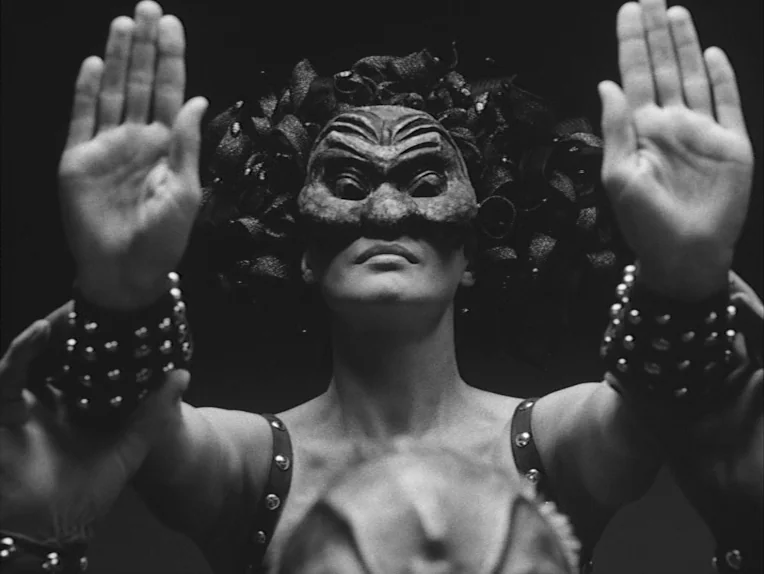 The Rite (1969) movie still: closeup — A performer stands before a black backdrop wearing a mask and studded leather accessories. They hold both…