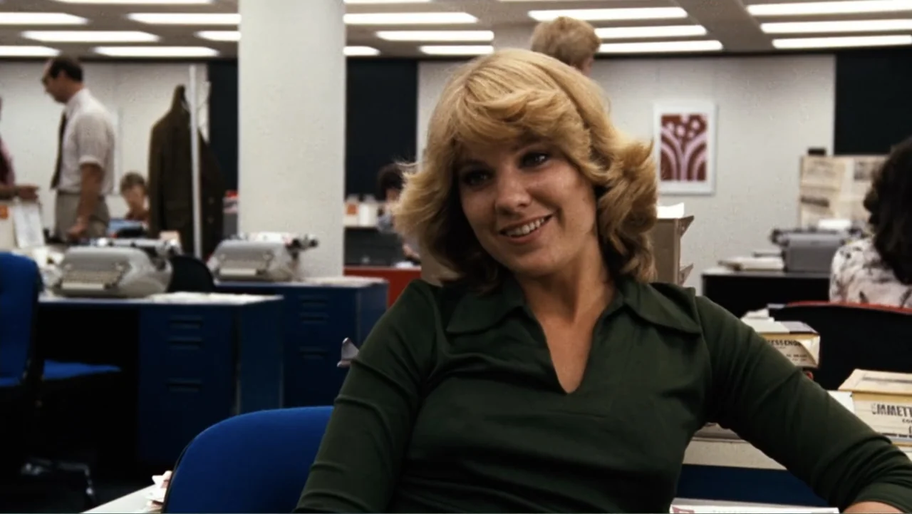 All the President's Men (1976) movie still: medium — A 1970s workplace environment with rows of blue desks and typewriter machines. A woman with…