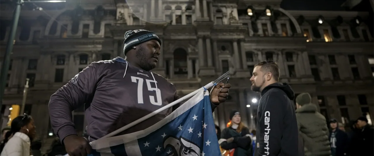 Kelce (2023) movie still: medium — A group of people gathers in front of a large, stone municipal building at night. A man in a sports jersey…