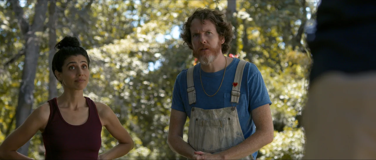Songs For A Sloth (2021) movie still: medium — A sunny outdoor park area features dense green foliage and trees in the background. Two individuals…