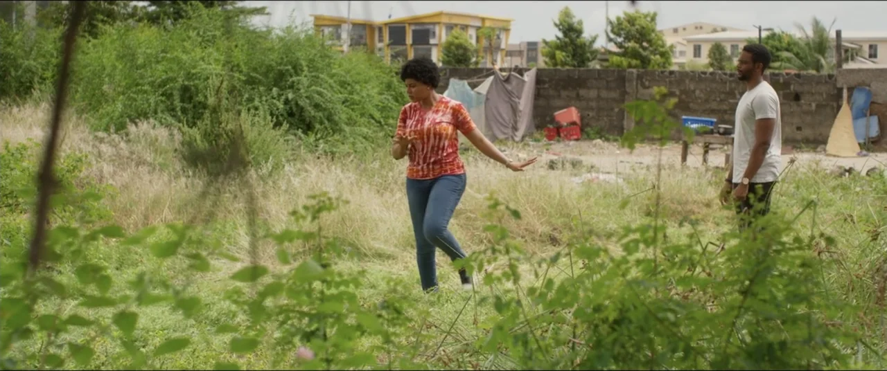 Saving Onome (2024) movie still: fullbody — An overgrown vacant lot with dry grass and a cinder block wall background. A woman in a tie-dye shirt…