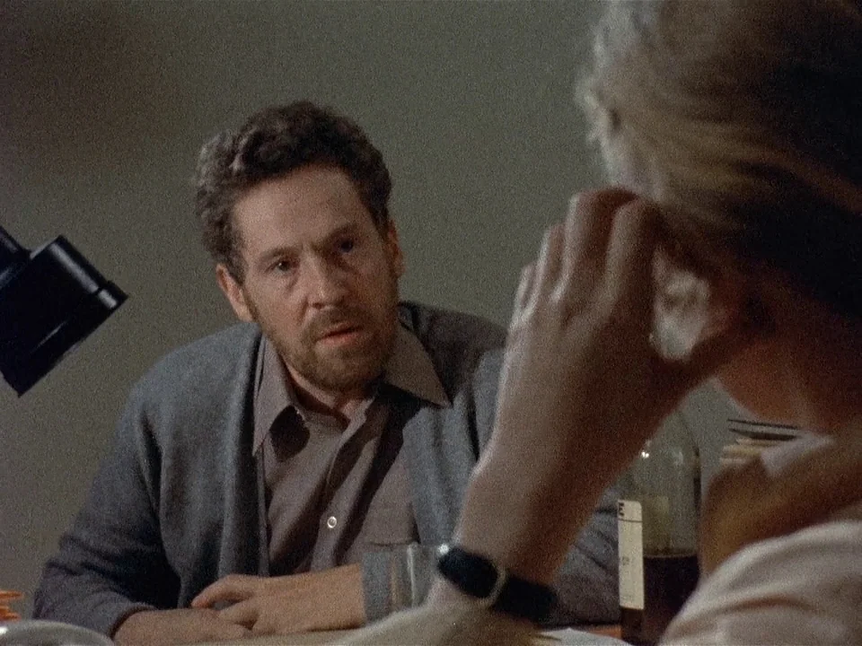 Scenes from a Marriage (1973) movie still: medium — A dimly lit room contains a desk where a man engages in conversation. The foreground shows the…