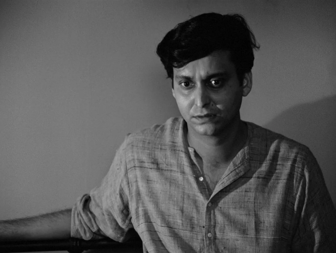 The Coward (1965) movie still: medium — A man sits against a plain, dimly lit wall in a sparse room. He looks directly at the camera with a subtle…