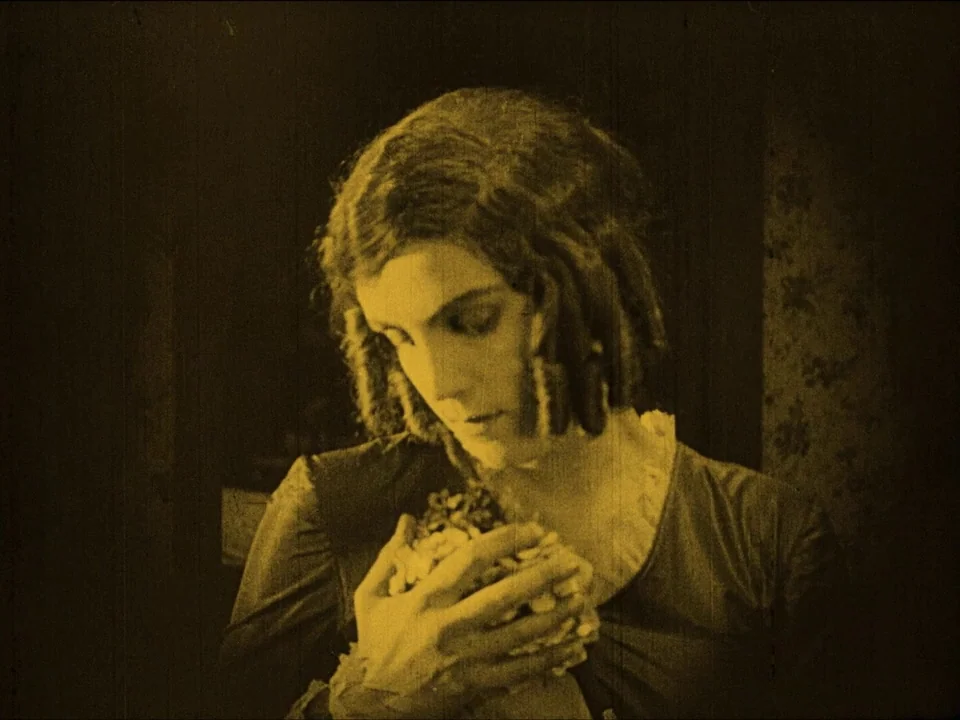 Nosferatu (1922) movie still: medium — A woman stands in a dark room with patterned wallpaper visible to the side. She looks downward while…