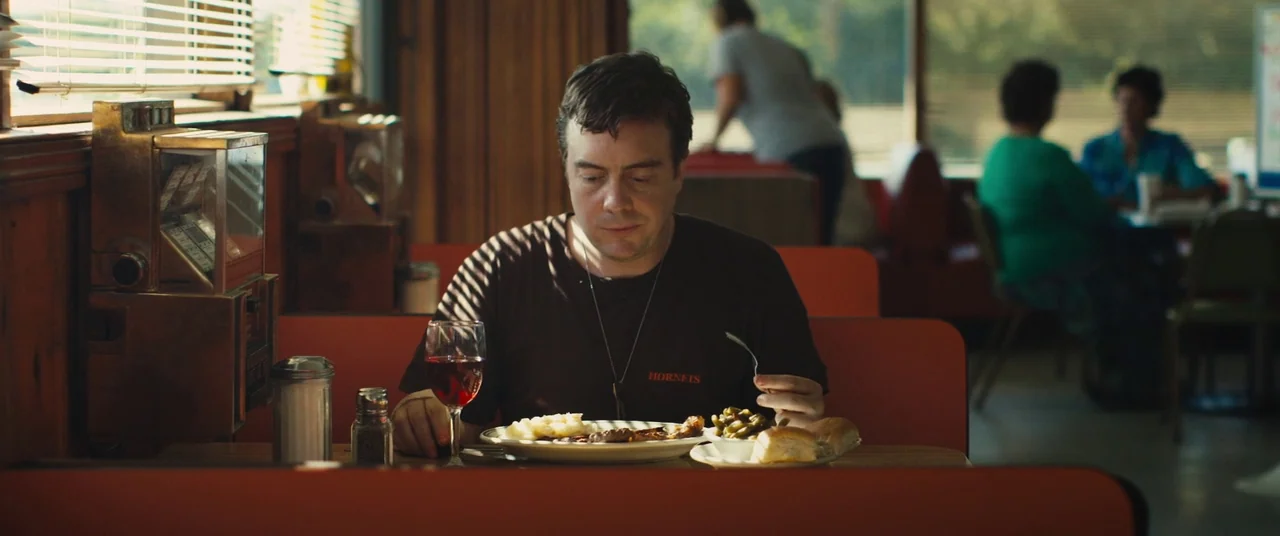 Blue Ruin (2013) movie still: medium — A diner booth features orange vinyl benches and wooden wall paneling with window blinds. A man sits alone at…