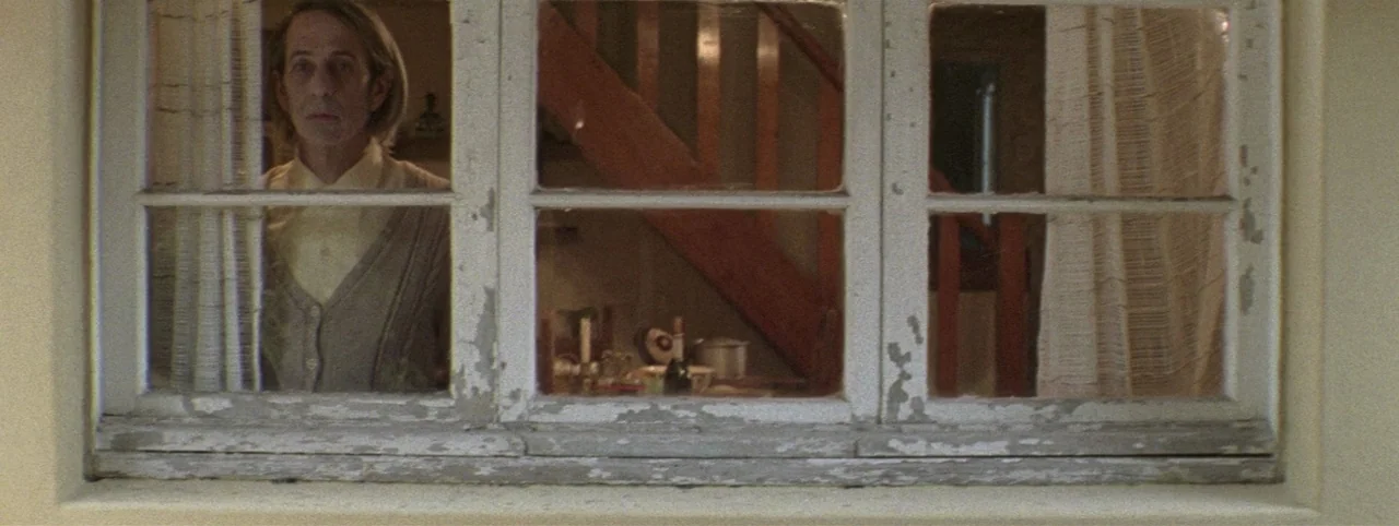 Post Mortem (2010) movie still: medium — A weathered white wooden window frame with peeling paint is set into a plain wall. A man in a grey cardigan…