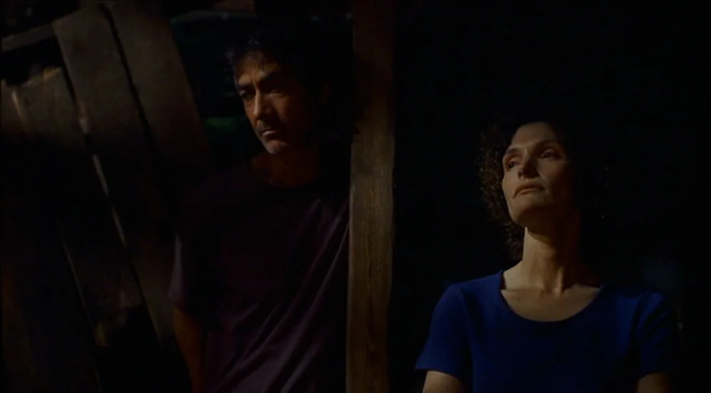 Limbo (1999) movie still: medium — A dark, minimally lit space features rough-hewn timber walls and beams. A man and a woman stand near a wooden…