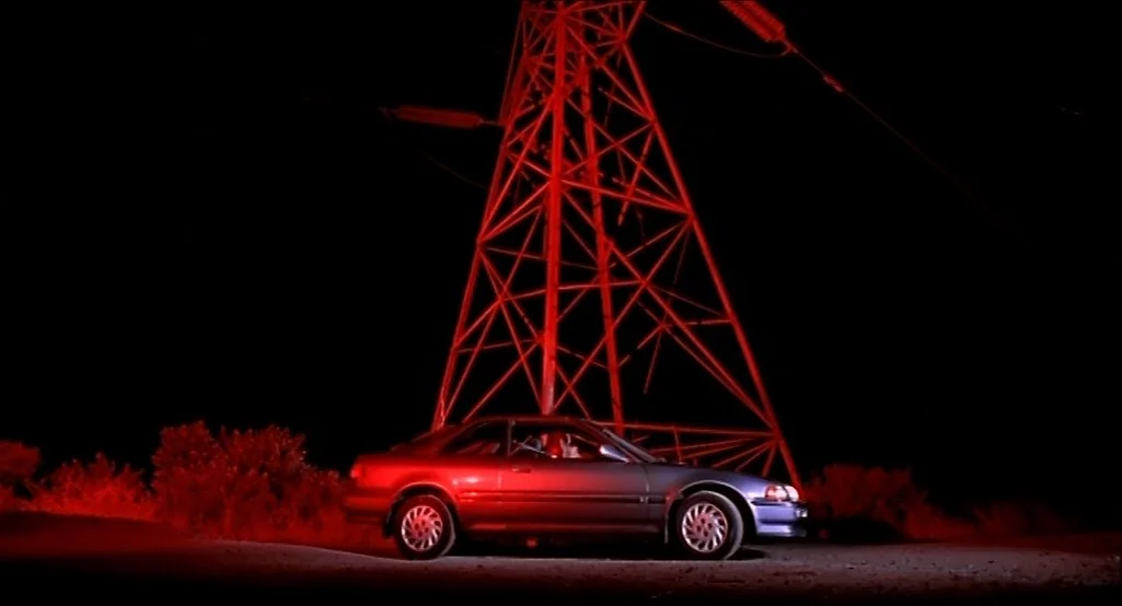 Nowhere (1997) movie still: wide — A dark, unpaved road at night features a sedan parked near a tall structure. The entire scene is bathed in a…