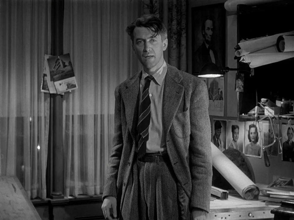 It’s A Wonderful Life (1946) movie still: medium — A dimly lit workspace with rolled architectural plans and photographs mounted on the wall. A man…