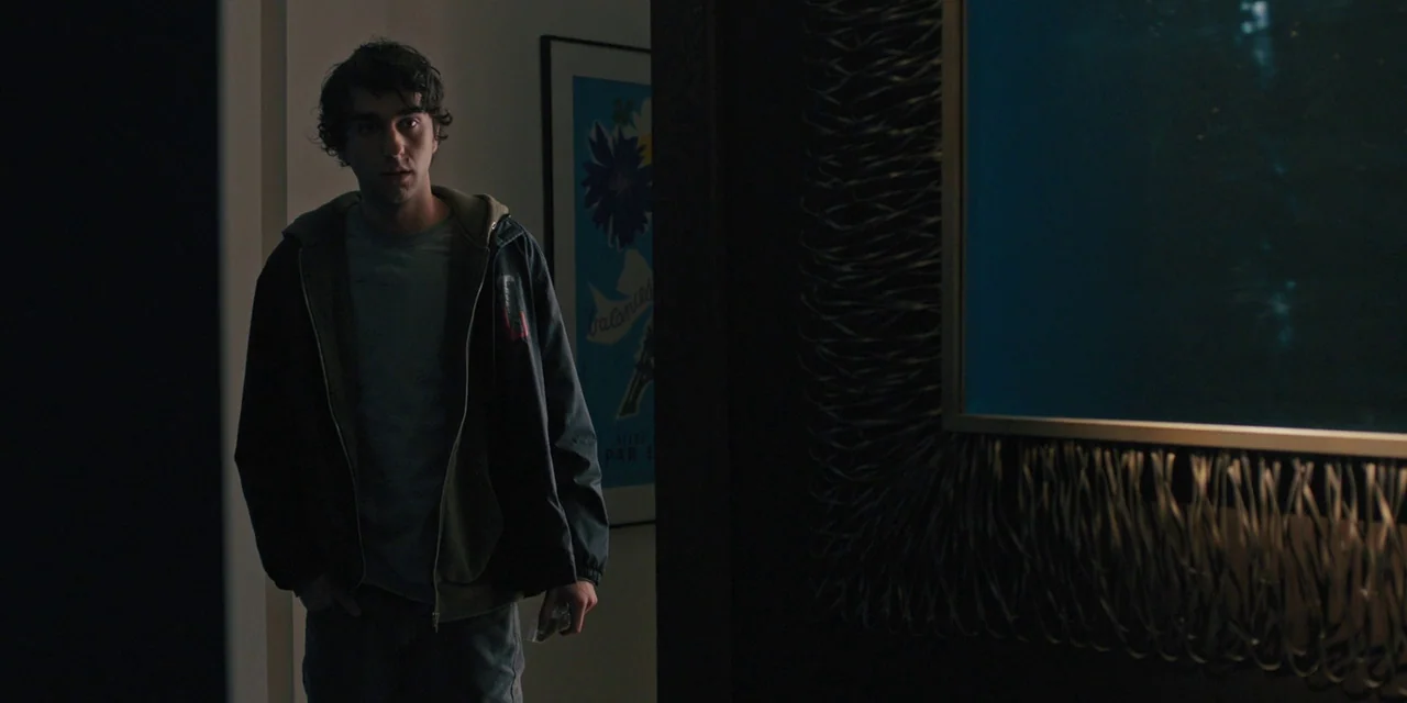 Hereditary (2018) movie still: medium — A dimly lit interior space features a textured dark frame on the right wall. A young man stands in the…