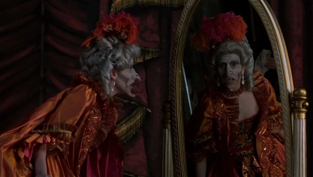 Interview With The Vampire movie still: medium — A dimly lit room features heavy dark red curtains draped in the background. An elderly woman in an…