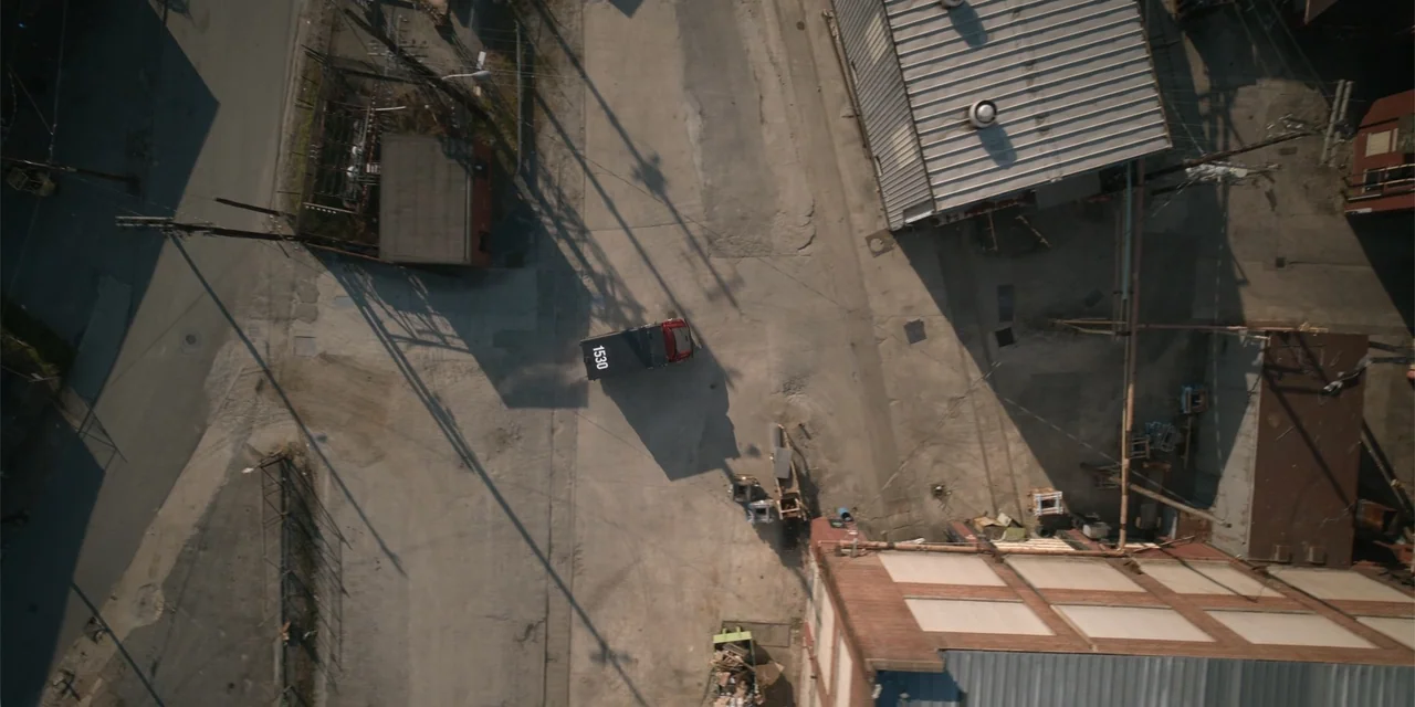 The Pickup (2025) movie still: establishing — An industrial paved lot is surrounded by brick warehouses and corrugated metal structures. A utility…