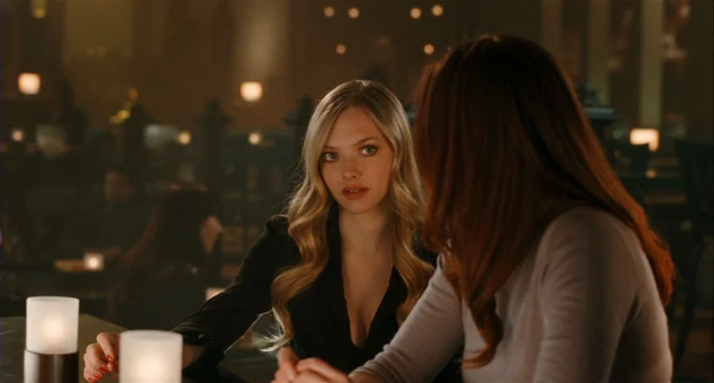 Chloe (2009) movie still: medium — A dimly lit bar interior features scattered candles on dark wooden surfaces at night. A blonde woman in a black…