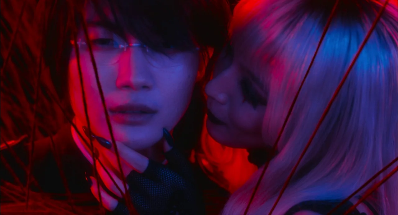 xxxHolic (2022) movie still: closeup — A close-up frame shows two individuals illuminated by intense red and blue light. Thin, dark branches cut…