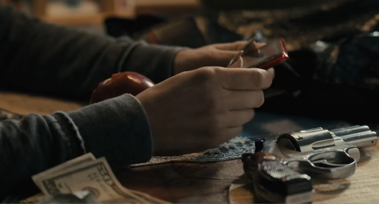 Lila & Eve (2015) movie still: closeup — A wooden table contains a revolver, cash, an apple, and a smartphone. Hands rest on the table surface while…
