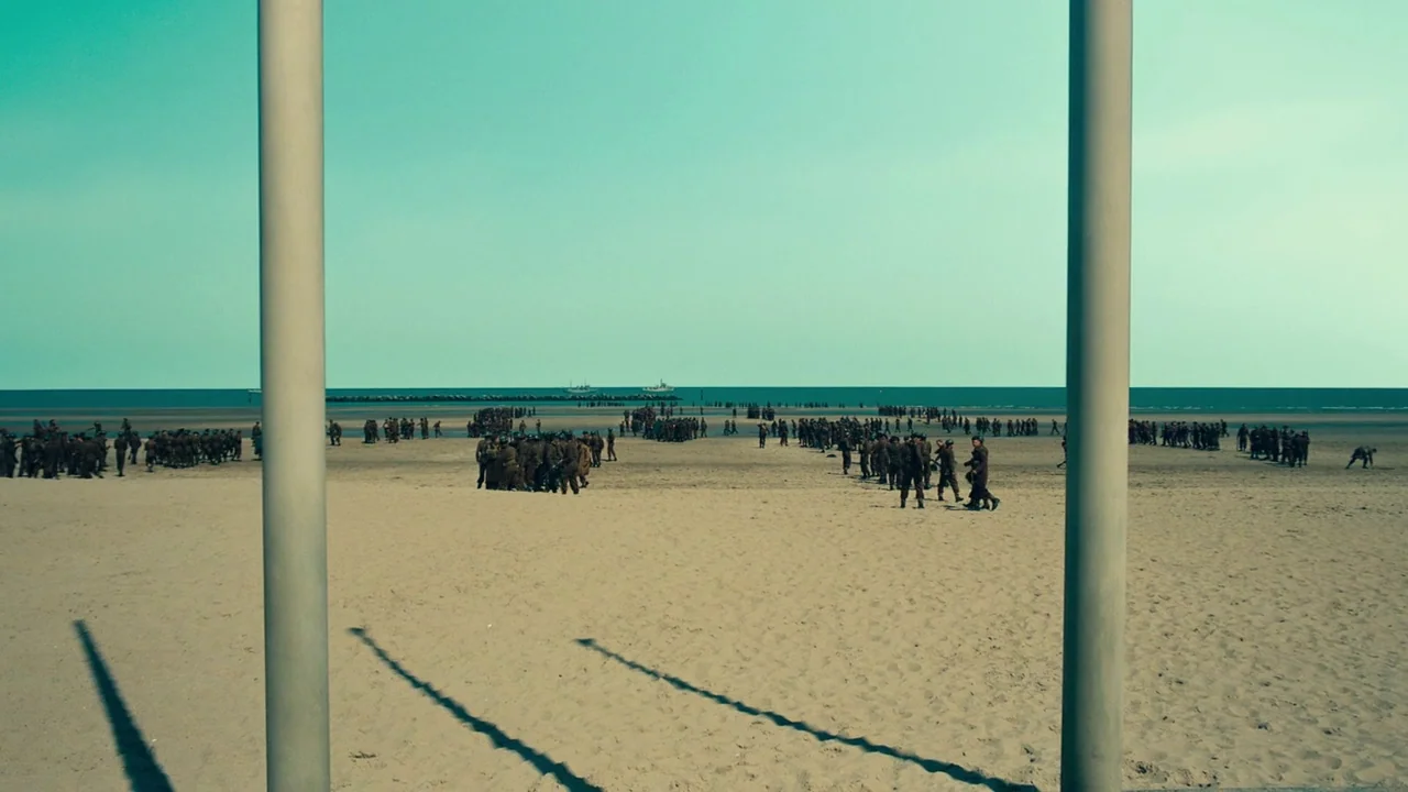 Dunkirk (2017) movie still: wide — A wide, flat sandy beach stretches toward a distant ocean horizon under a clear sky. Numerous soldiers in…