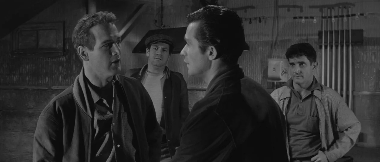 The Hustler (1961) movie still: medium — A dimly lit room with walls featuring textured metal paneling. Four men stand in a group, facing each other…