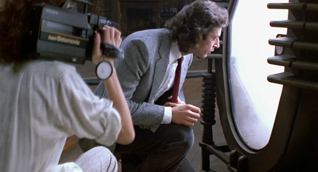 The Fly (1986) movie still: medium — A dimly lit room features metallic technology and wooden flooring. A man in a blazer examines a brightly…