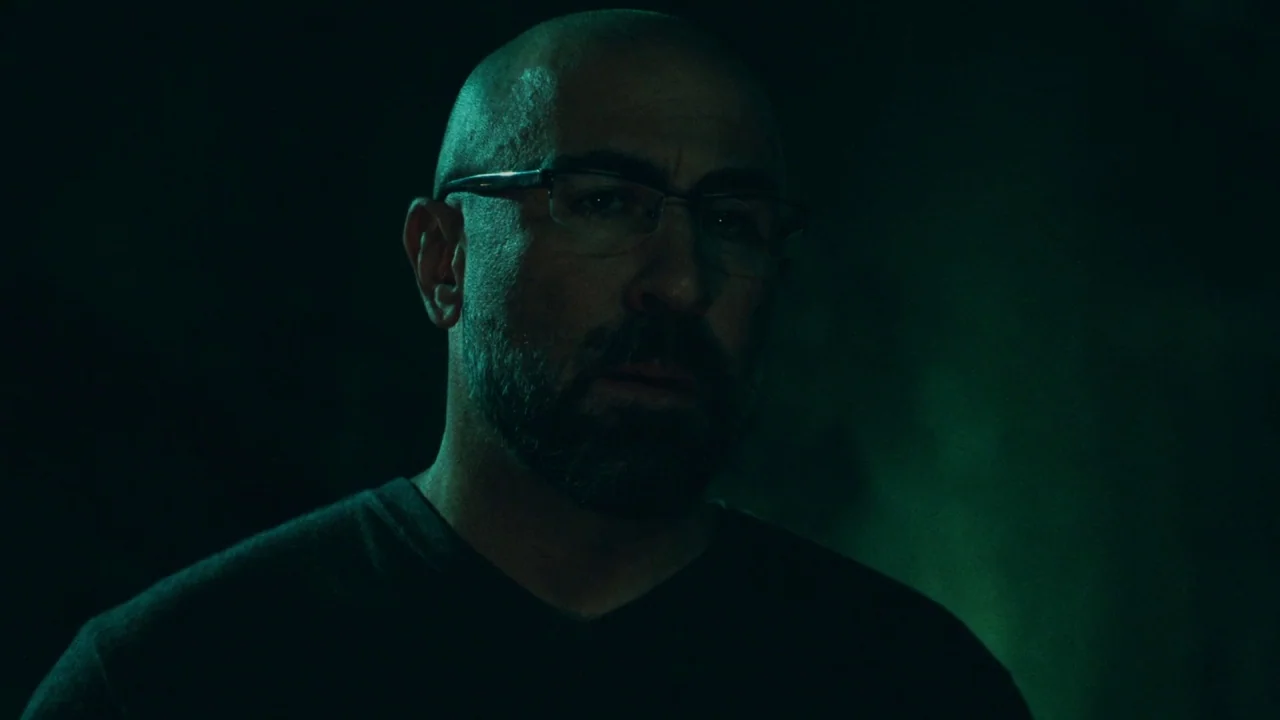 Saw V (2008) movie still: closeup — The subject stands in a darkened space illuminated by a dim green light. The man looks forward with a neutral…