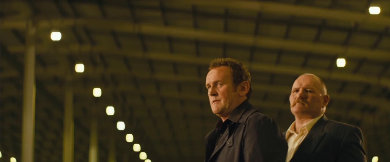 Layer Cake (2004) movie still: medium — A dark, industrial parking structure is illuminated by rows of overhead fluorescent lights at night. Two men…