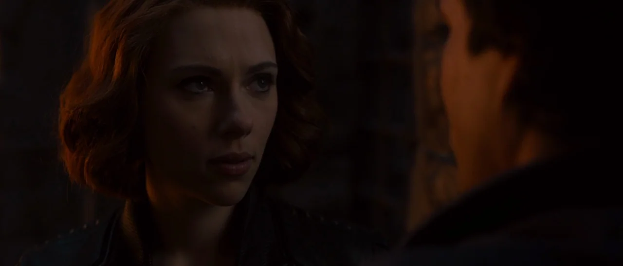 Avengers: Age of Ultron (2015) movie still: closeup — A dimly lit interior space with dark, textured backgrounds. A woman with auburn hair looks…