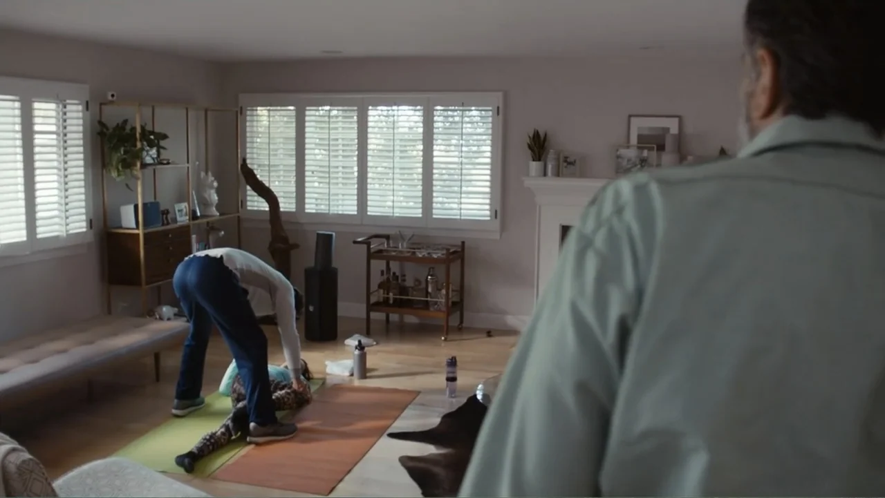 The Cleaner (2021) movie still: medium — A well-lit contemporary living room contains a yoga mat, bookshelf, and bar cart. A man stands over a woman…