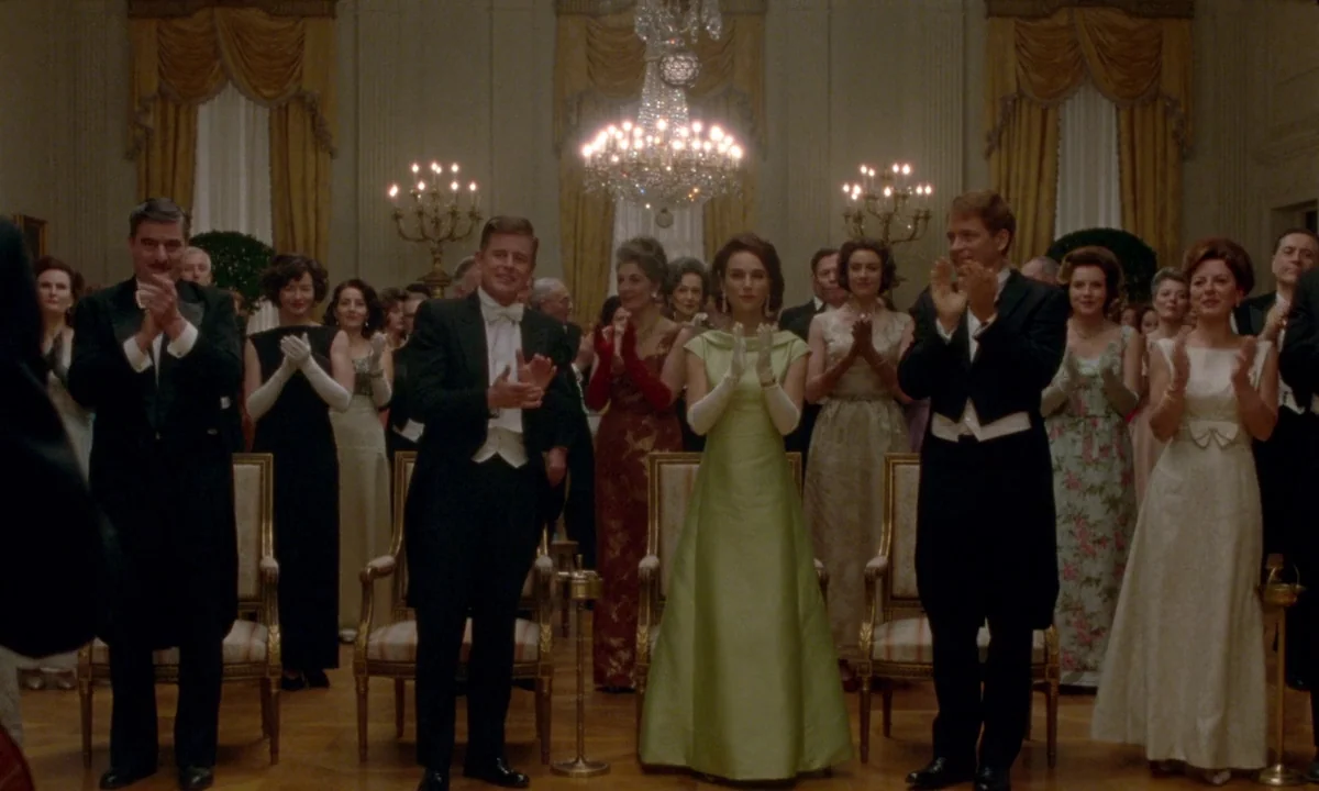 Jackie (2016) movie still: wide — An opulent 1960s ballroom features gold curtains, crystal chandeliers, and polished wooden floors. A large crowd…