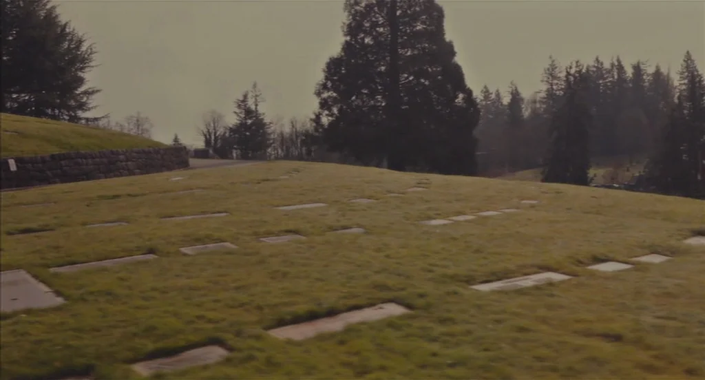 Restless (2011) movie still: establishing — A grassy hillside cemetery features rows of flat stone markers under a cloudy sky. Evergreen trees line…
