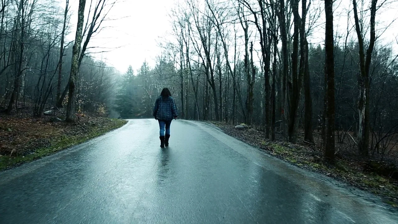 The Deeper You Dig (2019) movie still: wide — A wet asphalt road winds through a dense, leafless forest under an overcast sky. A lone woman wearing…
