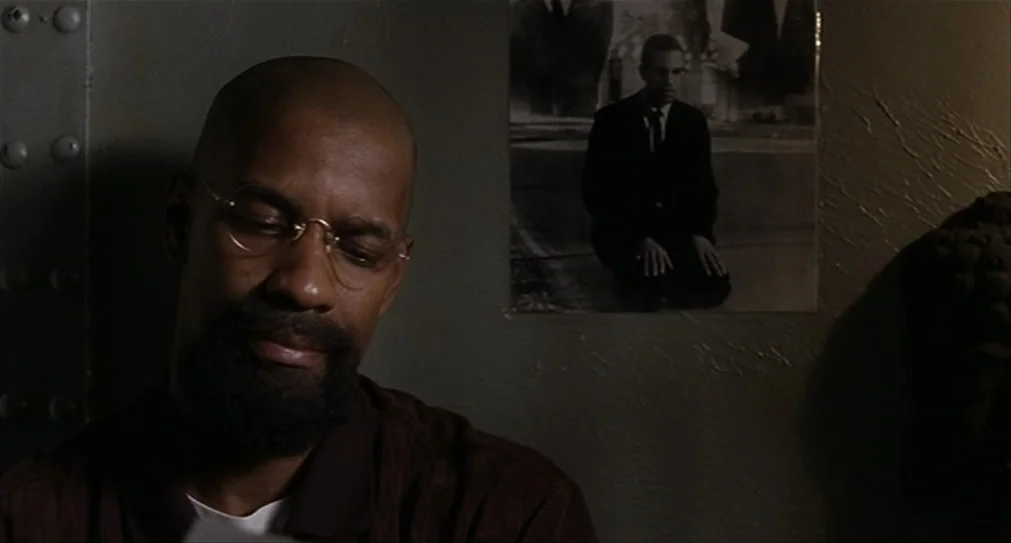 The Hurricane (1999) movie still: closeup — A dimly lit room with riveted steel walls features a poster of a man praying. A man with a beard and…