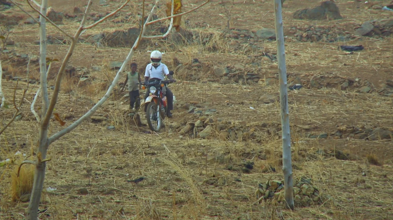 The Money Stone (2018) movie still: wide — A dry, dusty landscape filled with sparse brush and rocky ground patches. A man rides a motorbike past a…