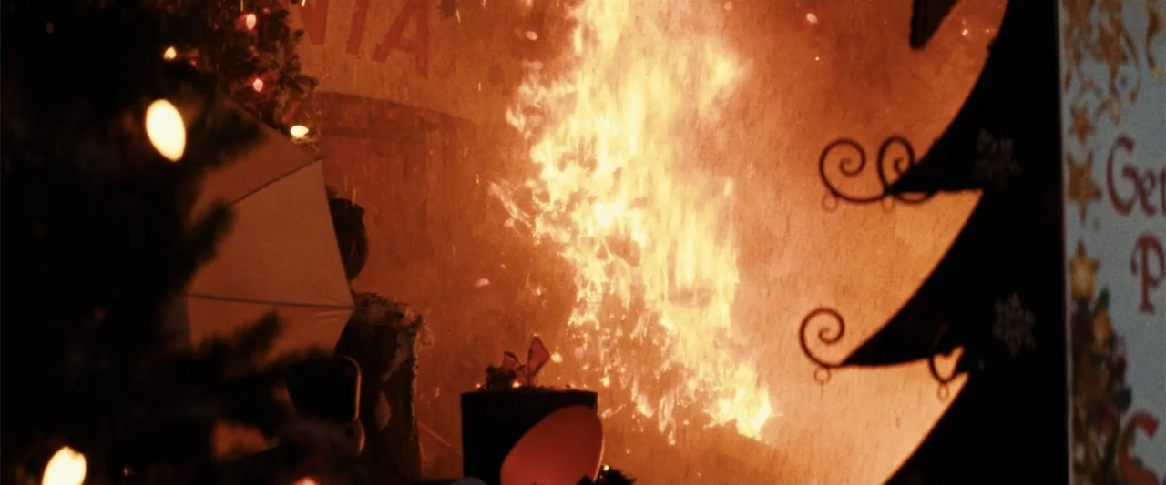 Terrifier 3 (2024) movie still: wide — An exterior storefront display features holiday decorations and a large, bright fire burning intensely. The…
