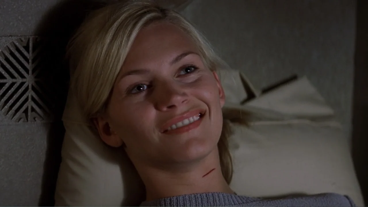 Ghosts of Mars (2001) movie still: closeup — A woman rests on a bed with a beige pillow and textured wall behind her. She smiles while looking…