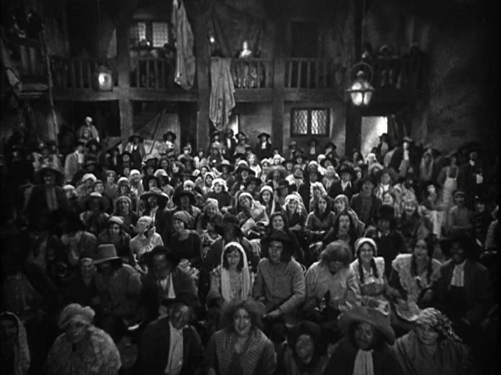 The Man Who Laughs (1928) movie still: wide — A dense crowd of people sits in an dimly lit, old-fashioned tiered theater space. The audience faces…