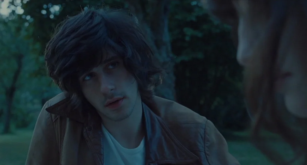 Something in the Air (2012) movie still: closeup — A man with messy dark hair sits outdoors during the evening blue hour. He looks toward someone…