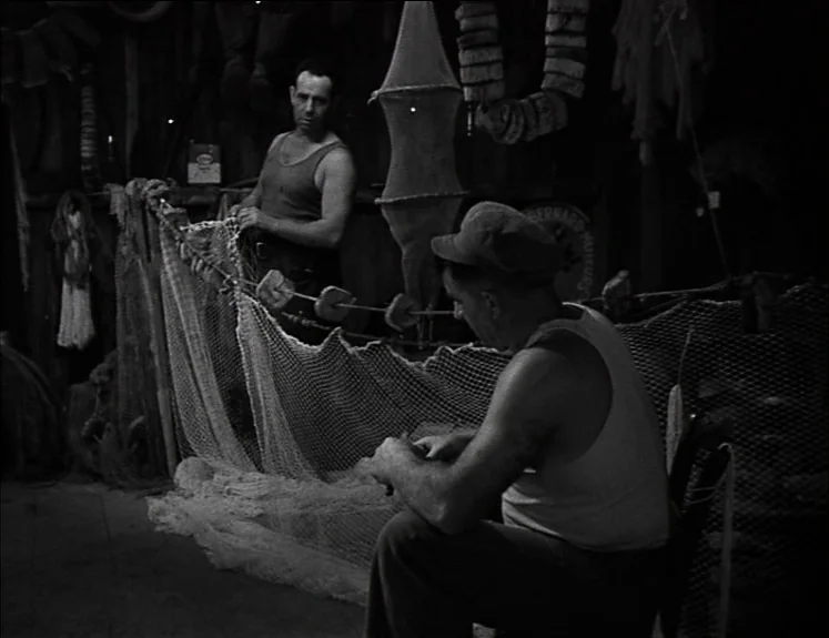 La Pointe Courte (1955) movie still: medium — A dimly lit boathouse contains various fishing equipment hanging from the wooden walls. Two men in…