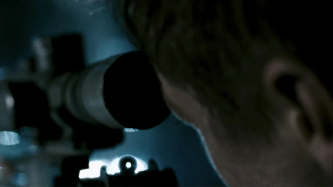28 Weeks Later (2007) movie still: closeup — A dark indoor space contains a person looking through a long tactical rifle scope. The person is in the…