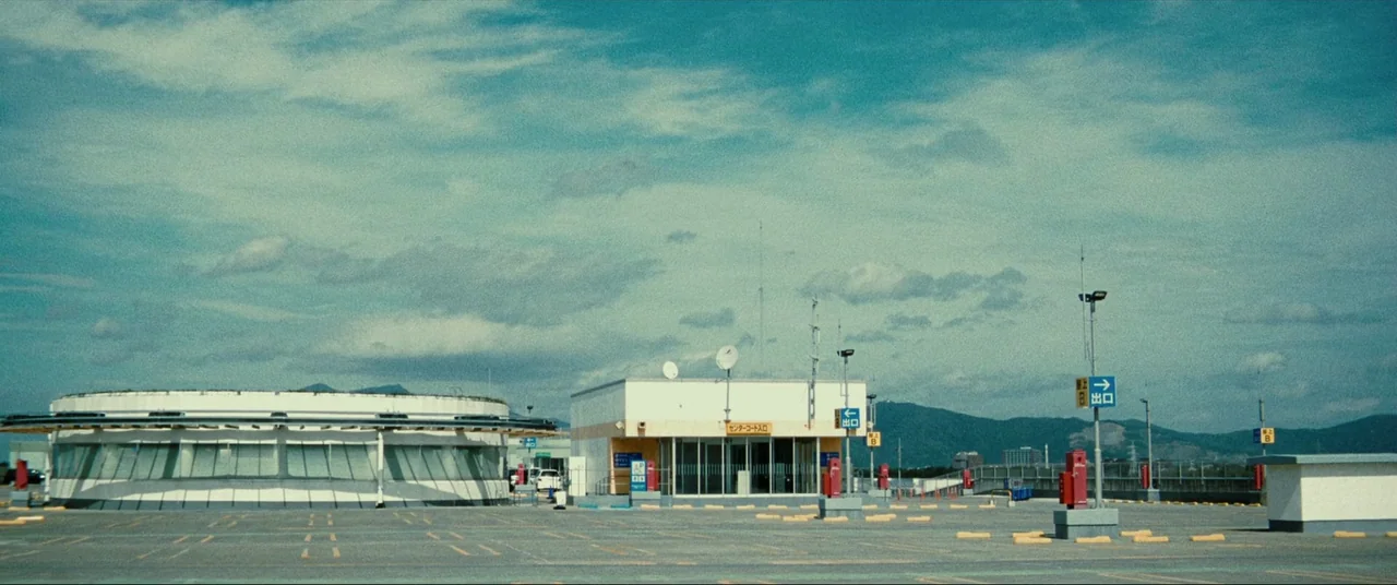 The World of Kanako (2014) movie still: establishing — A wide view of an empty asphalt parking lot featuring a circular white building. Low-rise…
