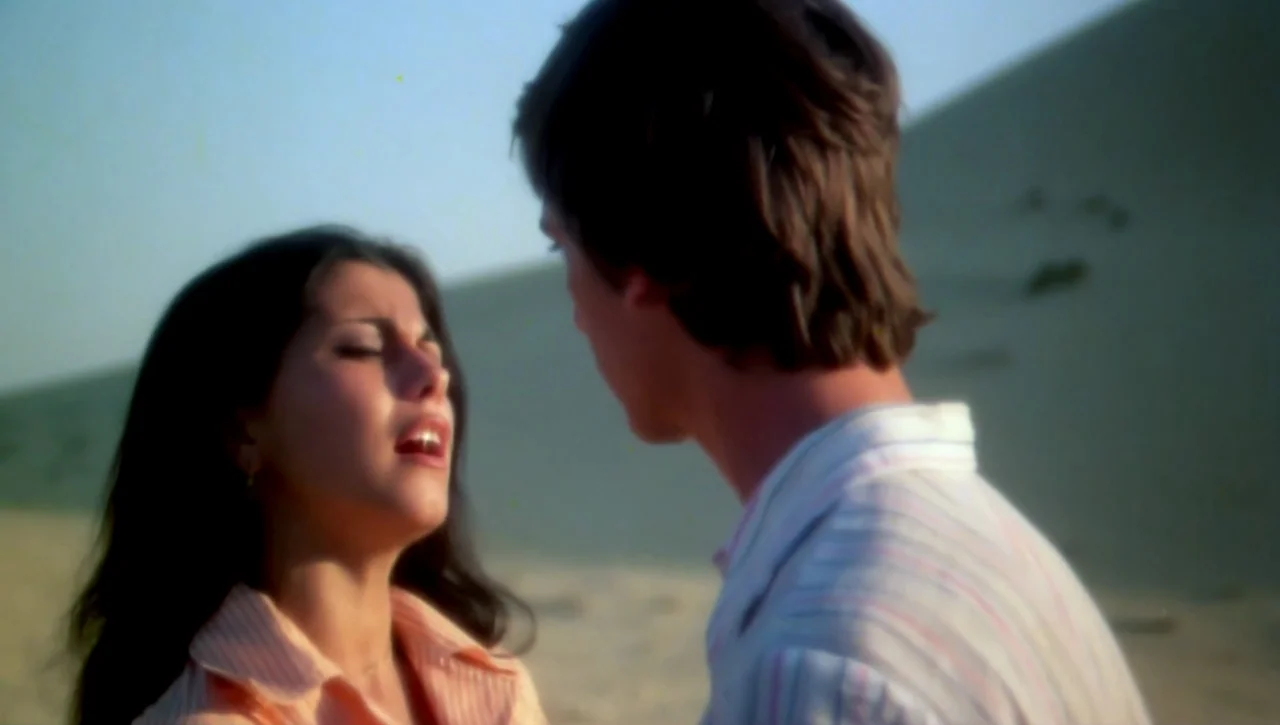 The Return of the Prodigal Son (1978) movie still: medium — A man and woman interact on a large, pale sand dune. The sky is clear during daylight…