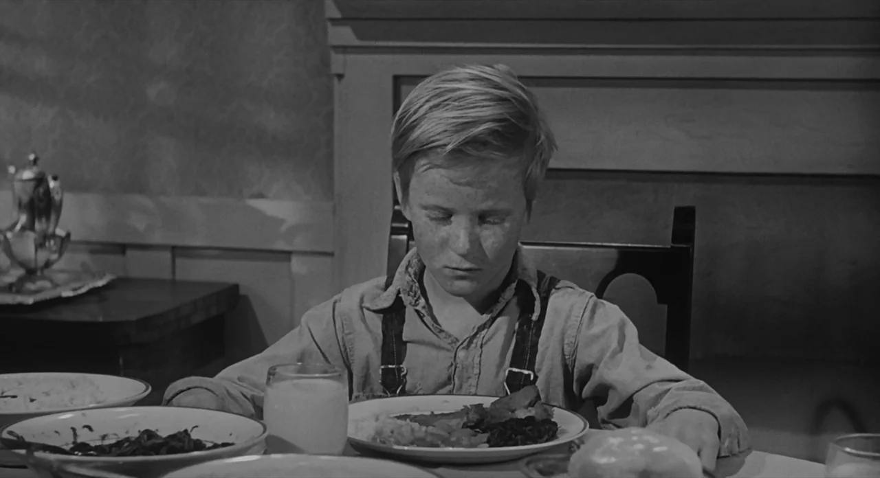 To Kill A Mockingbird (1962) movie still: medium — A rustic interior scene featuring a wooden table set for a meal. A young boy sits at the table…