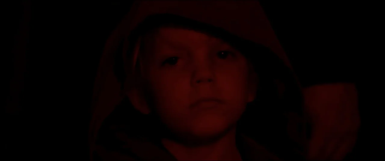 Amityville Cult (2021) movie still: closeup — A dark, unidentifiable space where the background is completely lost in shadows. A young child with a…