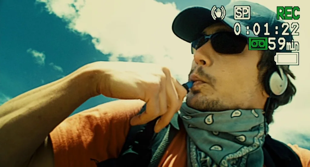 127 Hours (2010) movie still: closeup — A man in a baseball cap stands under a bright blue sky. He drinks from a plastic hydration tube while…