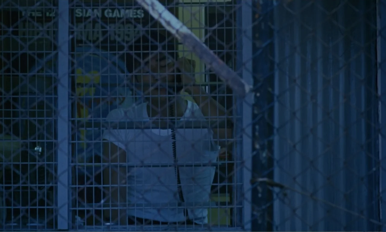 Chungking Express (1994) movie still: medium — A man stands inside a glass phone booth surrounded by a wire mesh fence. The man uses a corded…