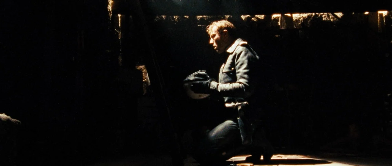 Let The Corpses Tan (2017) movie still: medium — A dark, cramped military bunker with rough, textured walls illuminated by a single light. A soldier…